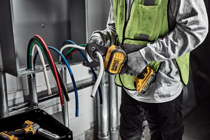 A person wearing gloves and a safety vest is using a DEWALT cordless cable cutter to cut a thick white electrical cable. Several colored cables are visible coming out of an electrical panel.