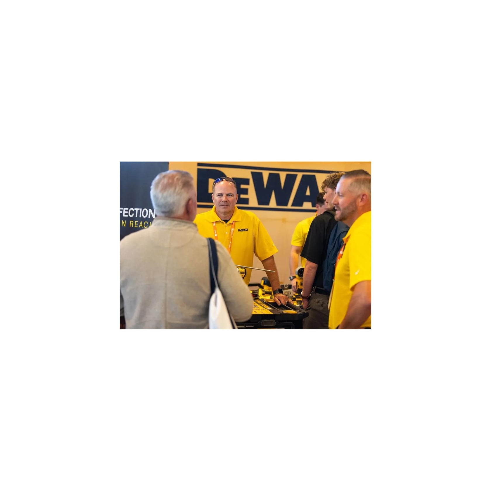 A group of people at a trade show booth observe a person in a yellow shirt demonstrating a DeWalt power tool with a bright light. Branded promotional items, tools, and a coffee cup are visible on the table.
