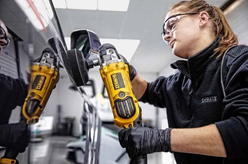 A person wearing black gloves and a DEWALT jacket is using a yellow DEWALT DWP849X polisher on the side of a car, with the reflection of the tool visible in the car's surface. The person's face is blurred.