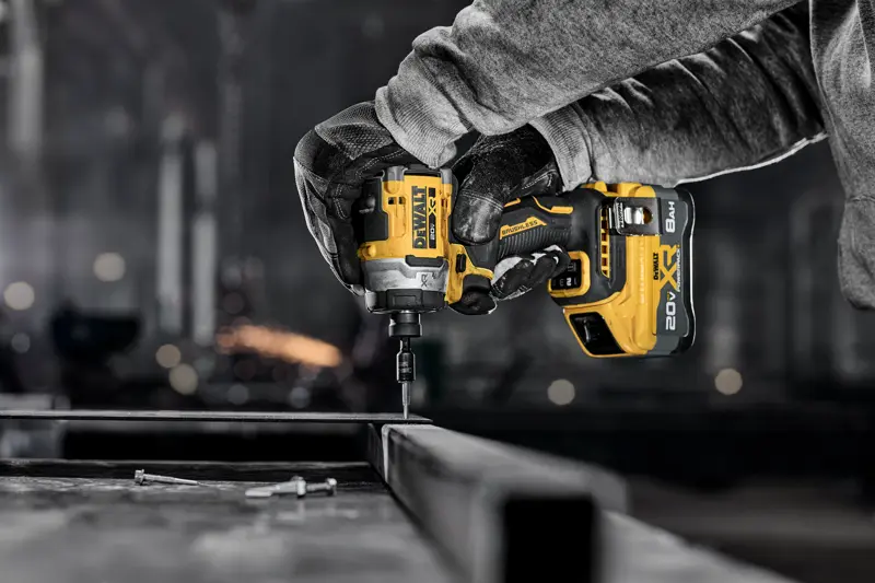 A person uses a DEWALT cordless impact driver to fasten a screw into metal in an industrial workshop setting.