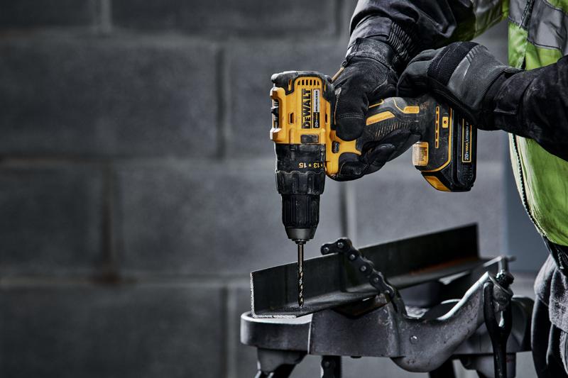 A close-up of a person using a DEWALT DCD777C2 cordless drill to drill into a metal beam in an industrial setting. The person is wearing gloves and a work jacket.