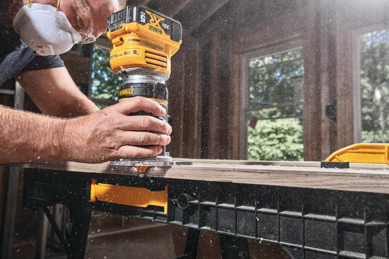 A person wearing a dust mask uses a yellow DEWALT cordless router on a piece of wood, creating dust in a workshop environment.