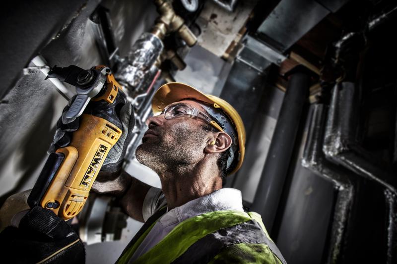 A person wearing a yellow hard hat and safety vest uses a DEWALT DWE315 power tool to cut or grind material on a wall in an industrial setting. Their face is obscured.