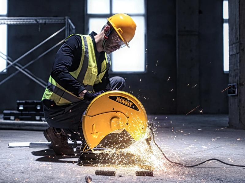 A person wearing a yellow safety helmet and reflective vest is using a DEWALT D28730 cutting tool to cut metal pipes in an industrial setting, with visible sparks flying from the machine.