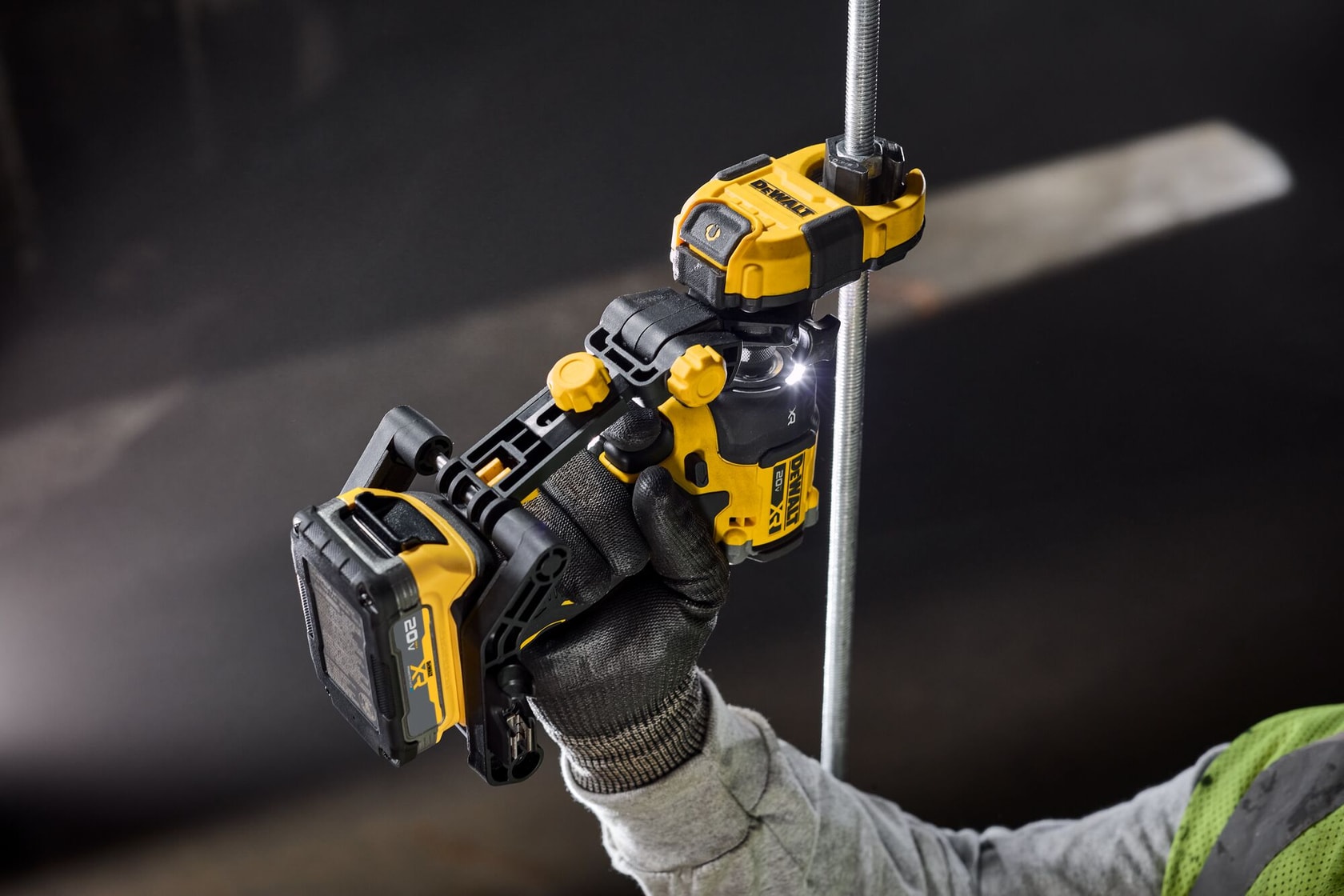 A close-up of a person holding a yellow and black DEWALT cordless power tool, attaching it to a vertical metal rod. The background is dark and the person is wearing a gray sleeve and a black glove.