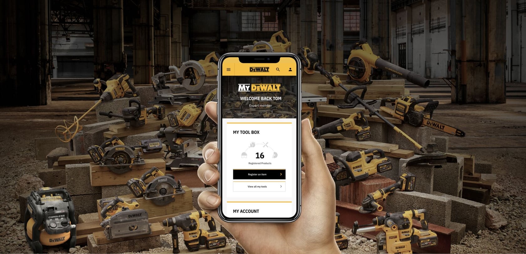 A hand holding a smartphone showing the My DEWALT account page, displaying registered products. In the background, there is a large assortment of DEWALT power tools arranged in an industrial workshop setting.