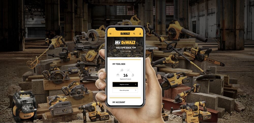 A hand holding a smartphone showing the My DEWALT account page, displaying registered products. In the background, there is a large assortment of DEWALT power tools arranged in an industrial workshop setting.