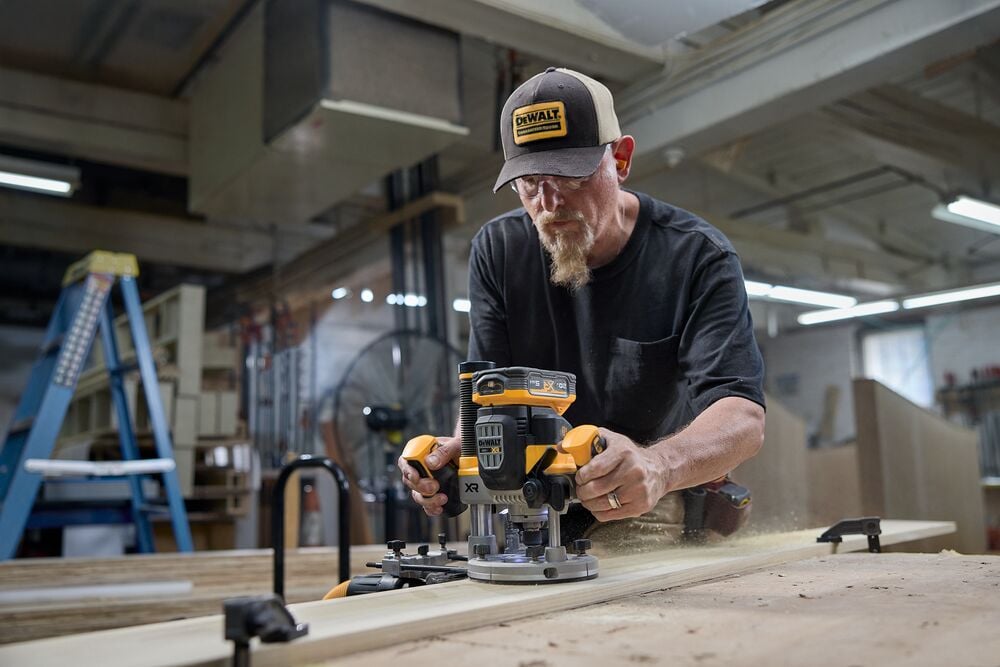 A person using a DEWALT cordless router to shape a piece of wood in a workshop. The person is wearing a DEWALT cap and working on a large wooden table, with sawdust visible and various workshop equipment in the background.