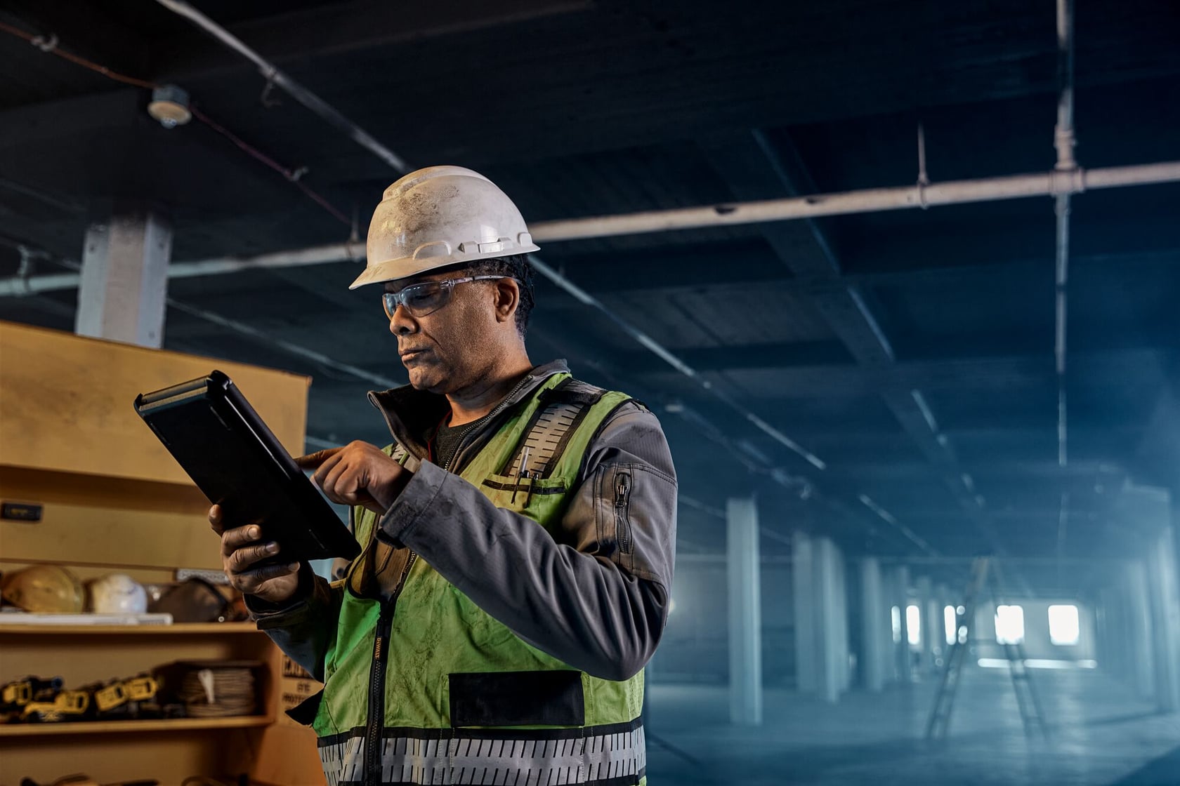 A construction worker wearing a hard hat and green safety vest uses a tablet device inside a large industrial building. Safety equipment is visible in the background.