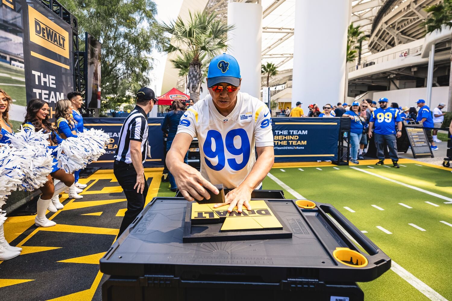 A person wearing a sports jersey with the number 99 and a blue cap participates in an outdoor event featuring DEWALT products. The scene is set on a football-themed area, with cheerleaders holding pompoms, a referee, and several people in similar jerseys in the background. The person is interacting with a DEWALT-branded black storage box.