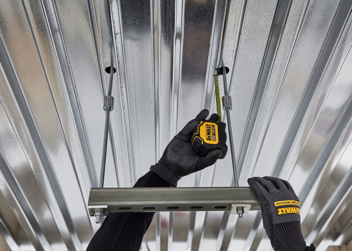 A person wearing black gloves with yellow DEWALT branding uses a yellow DEWALT tape measure to measure a metal rod suspended from a corrugated metal ceiling. The person holds a metal channel for support.