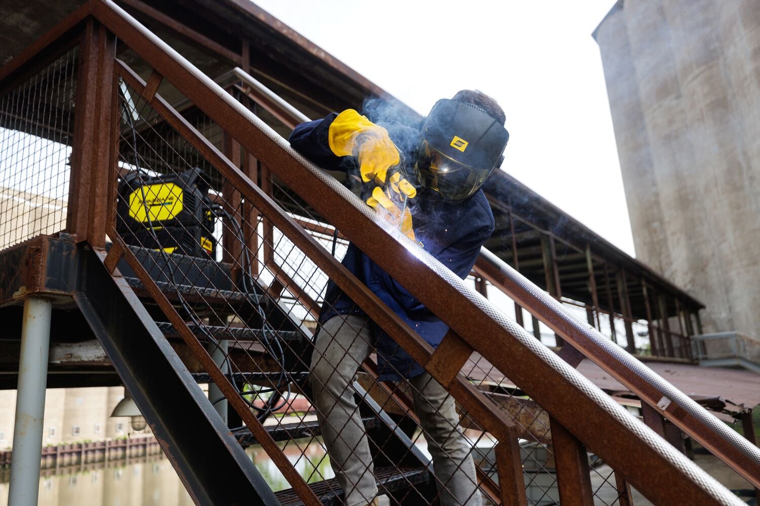A person wearing protective gear is welding a metal staircase outdoors. The individual has yellow gloves and a welding helmet, and there is visible smoke from the welding process. A DEWALT welding machine is positioned nearby on the steps.
