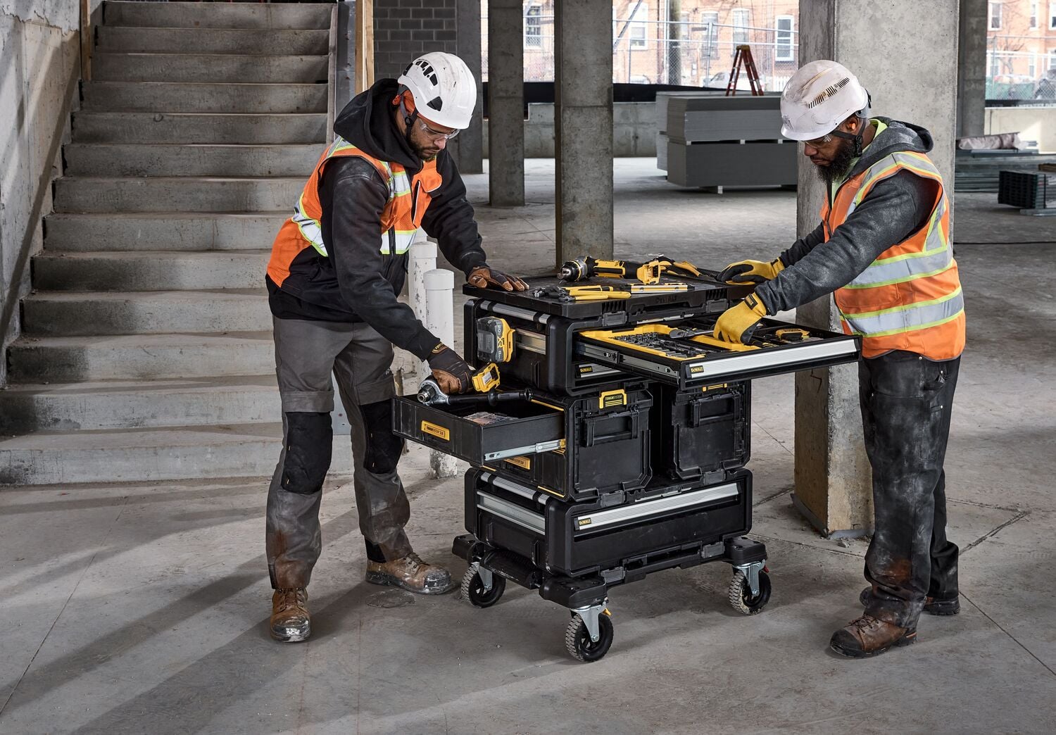 Two construction workers wearing safety gear and helmets are using a mobile DEWALT tool storage cart with multiple drawers and compartments, equipped with various power tools and accessories, inside an unfinished building.