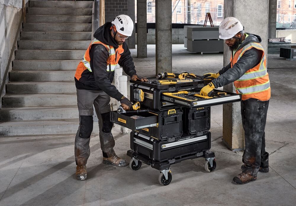 Two construction workers wearing safety vests and helmets are using a DEWALT rolling tool storage cart with multiple drawers and compartments in a partially finished building. Various DEWALT tools are placed on the cart.