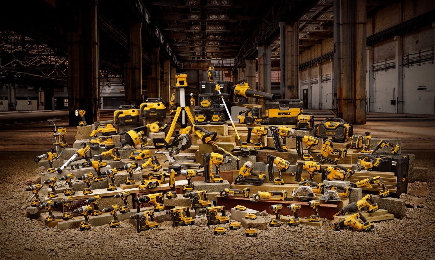 A large collection of DEWALT power tools, accessories, and toolboxes arranged on concrete blocks in an industrial warehouse setting. The tools include drills, saws, grinders, and other construction equipment, all in DEWALT's distinctive yellow and black colors.
