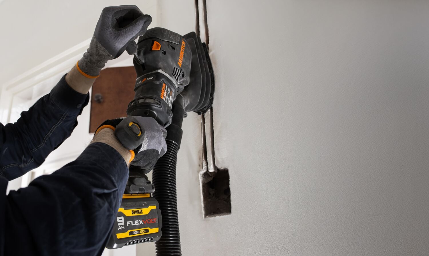 A person wearing gloves uses a DEWALT FLEXVOLT cordless power tool to sand or grind a section of a white wall next to an electrical outlet opening.