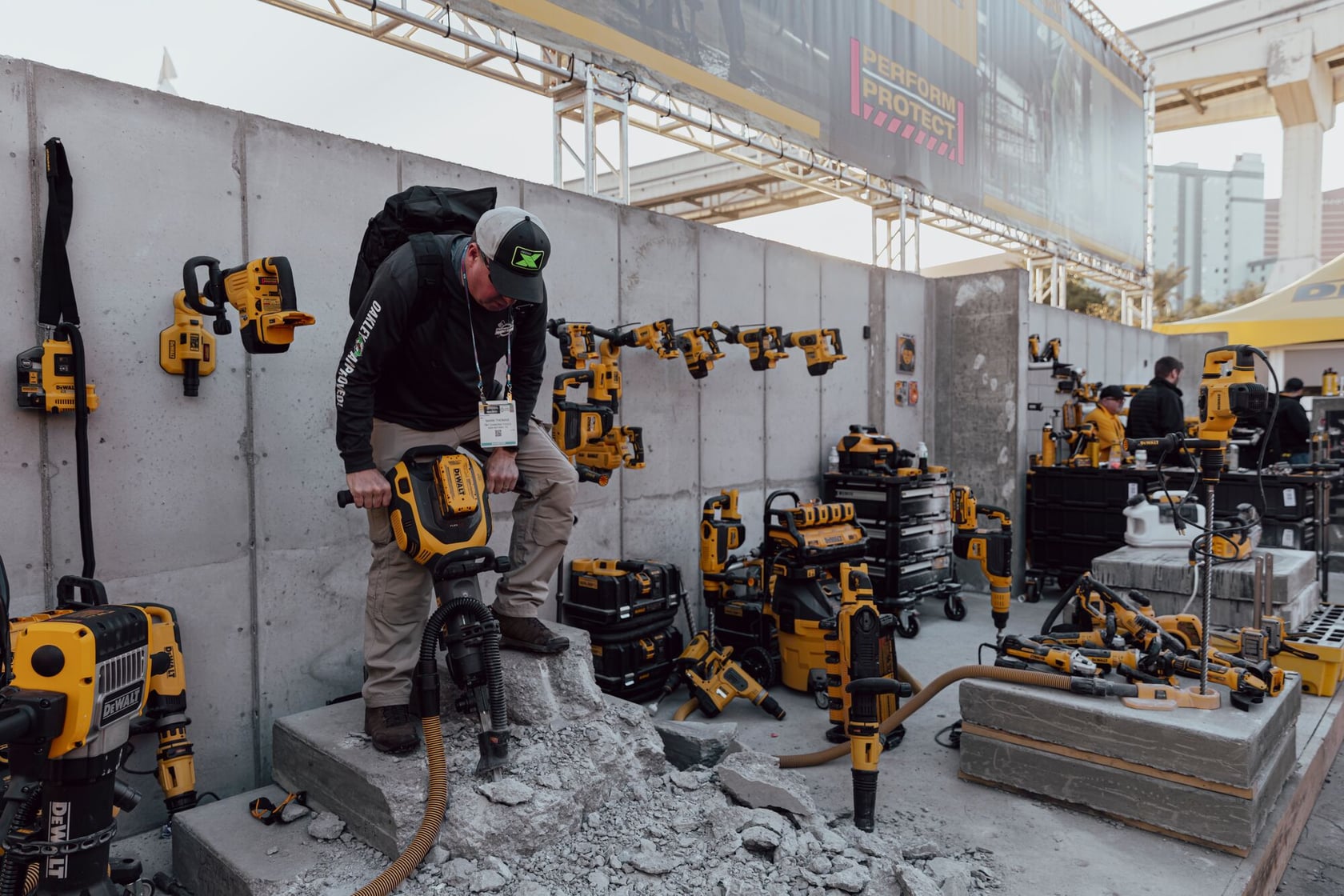 A person is using a yellow and black industrial jackhammer to break through concrete at a demonstration area. The background features multiple yellow and black power tools displayed on racks and walls, along with toolboxes and equipment, suggesting a product showcase or trade event.