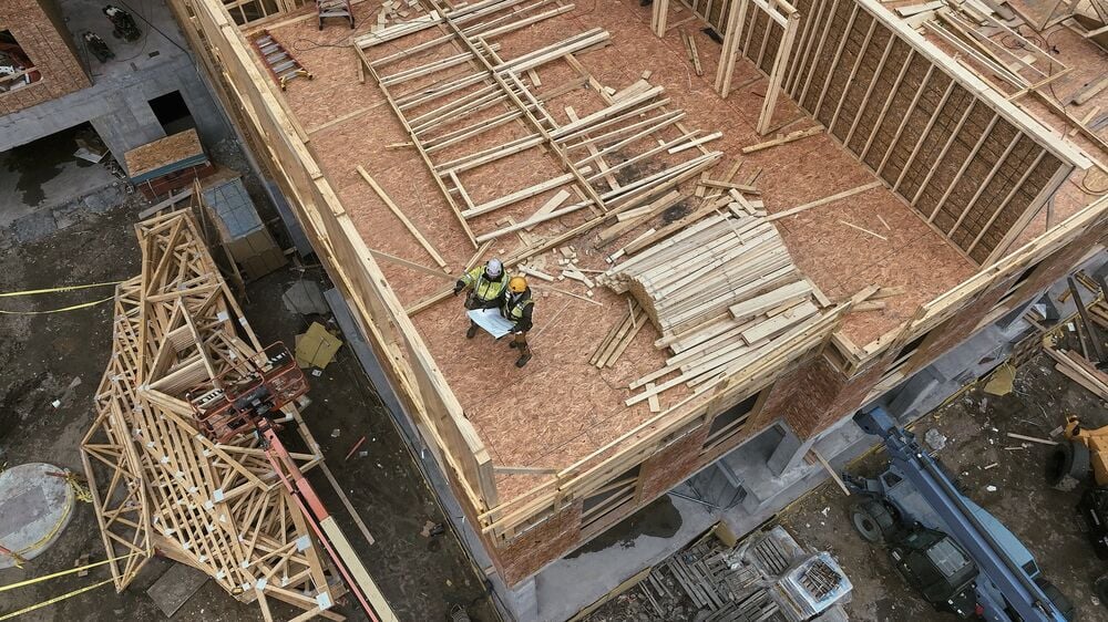 Aerial view of a construction site with two workers wearing safety gear standing on the wooden framework of a building. Various construction materials and equipment are visible around the site.
