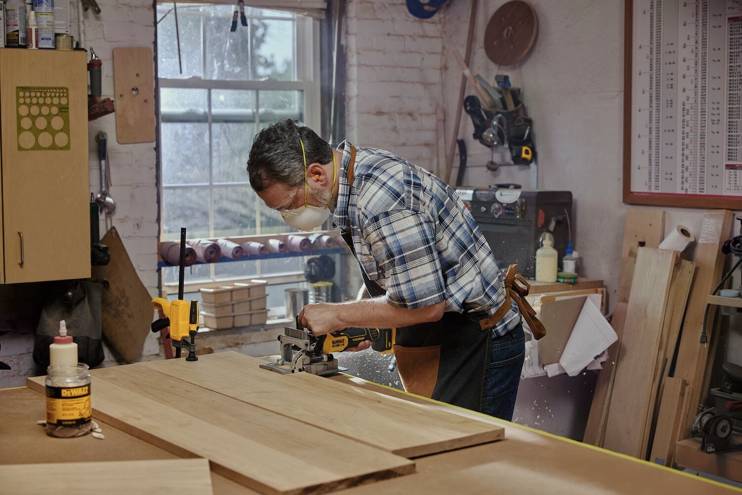 A person using a DEWALT power tool to work on wooden boards in a woodworking shop. The workspace contains various tools, equipment, and a bottle of DEWALT wood glue on the table.