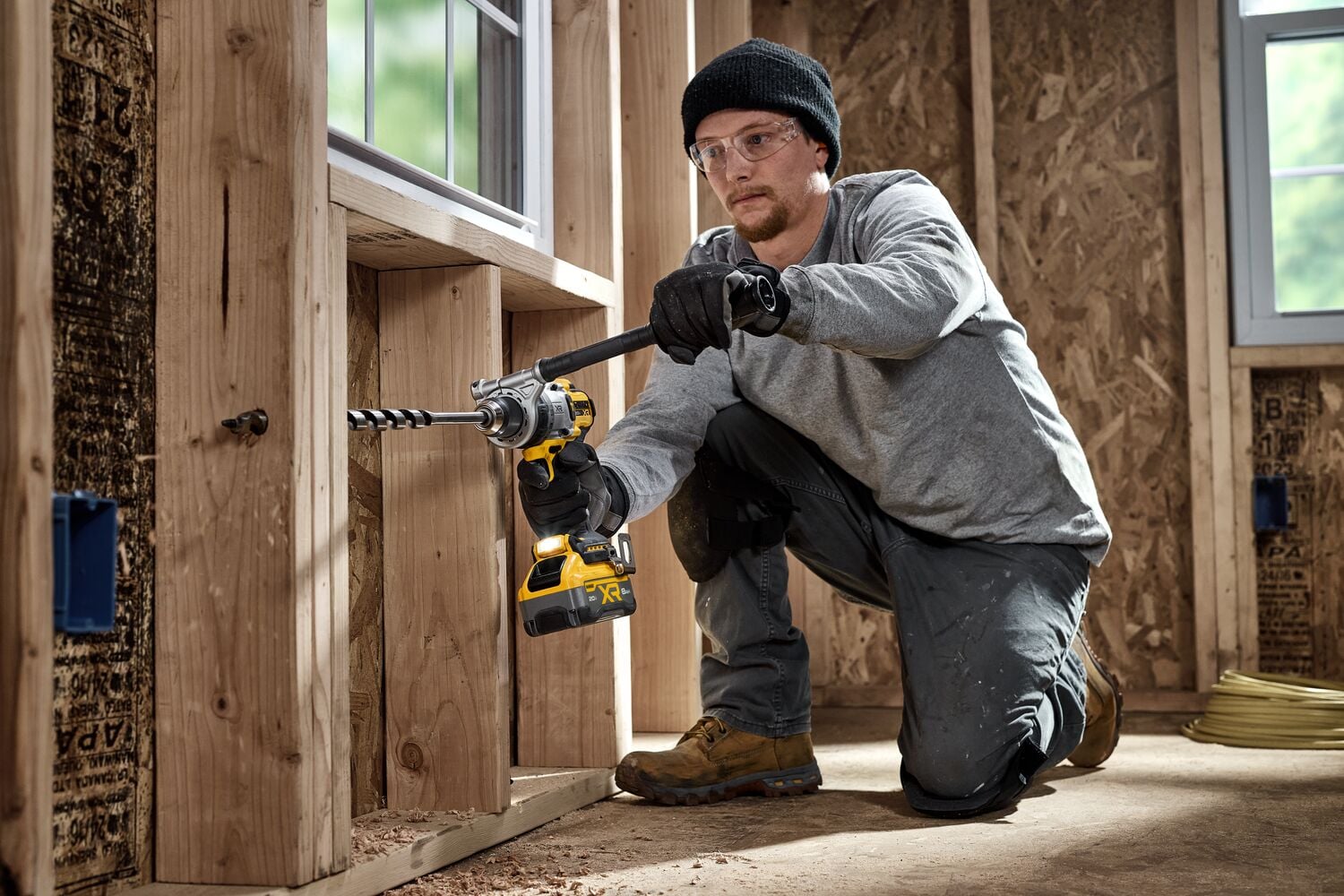 A person using a DEWALT cordless power drill to drill a hole into a wooden stud in a construction setting. The worker is kneeling and wearing work gloves, a hat, and boots. The background shows unfinished walls and windows.