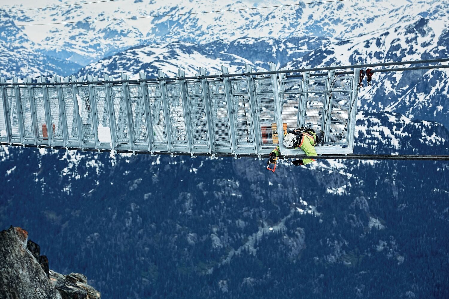 A construction worker wearing safety equipment is working on a metal suspension bridge high above a mountainous landscape with snow-covered peaks.