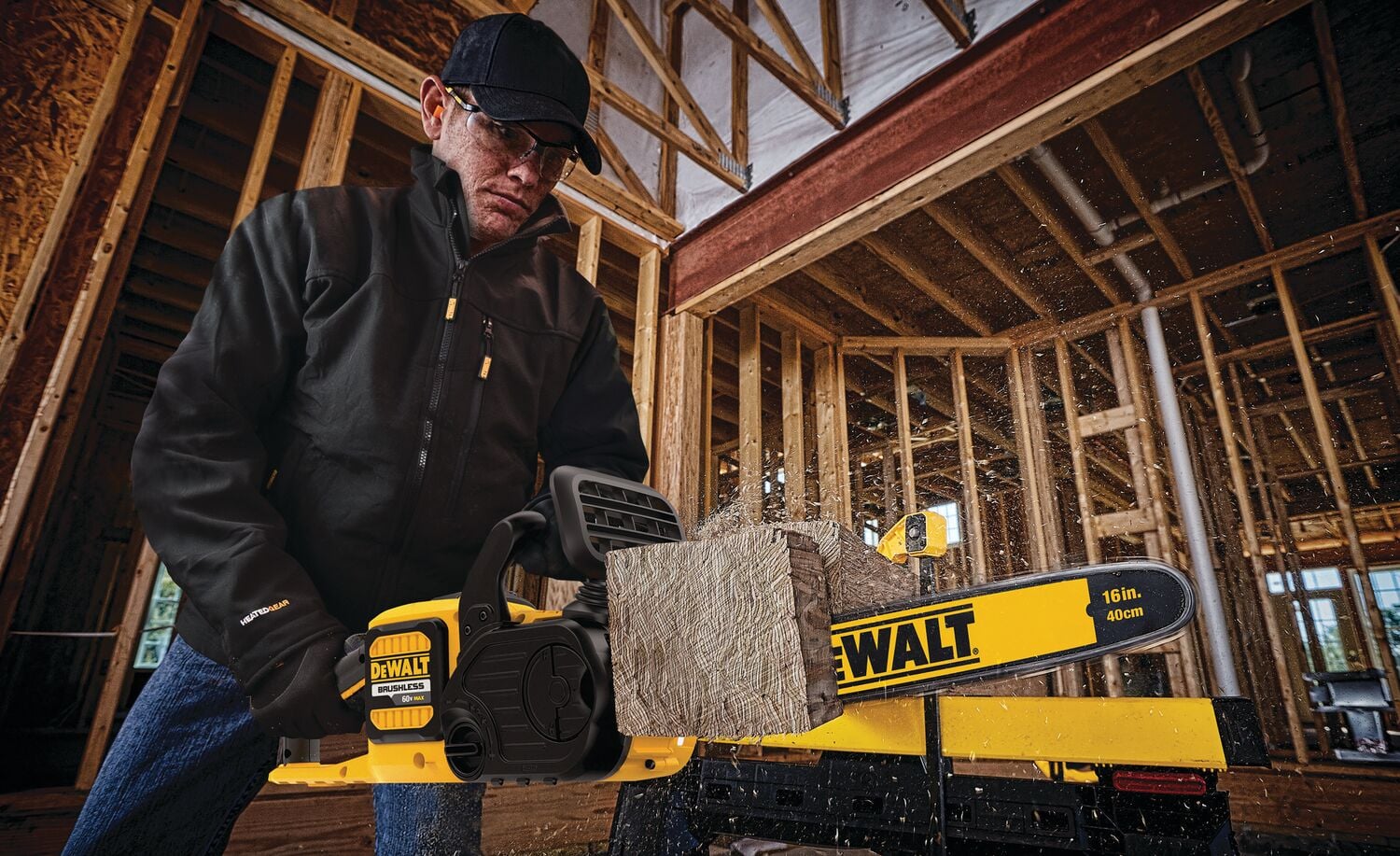 A person wearing a black jacket and gloves is using a yellow DEWALT chainsaw to cut a wooden beam inside a construction site with exposed framing.