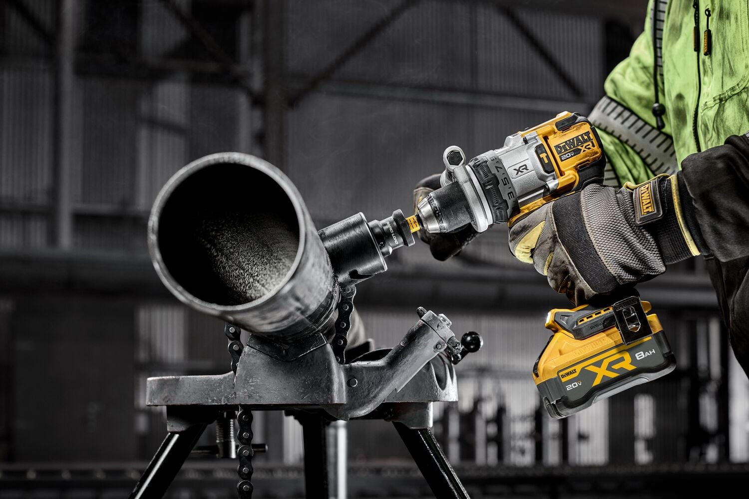 A person wearing gloves and a green jacket uses a DEWALT cordless drill to bore into a large metal pipe mounted on a stand in an industrial setting.