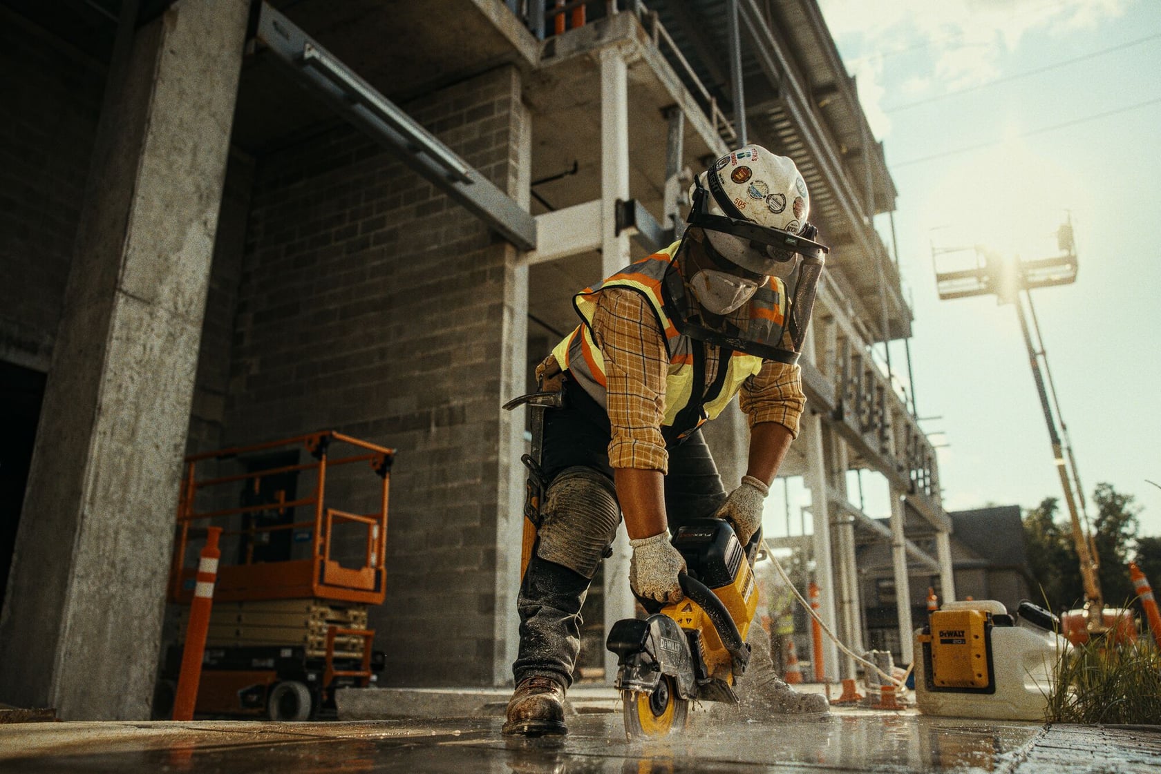 A construction worker wearing safety gear operates a DEWALT power saw to cut concrete at a construction site. The worker is using protective equipment such as a helmet, face shield, gloves, and reflective vest. The site includes unfinished building structures and equipment.