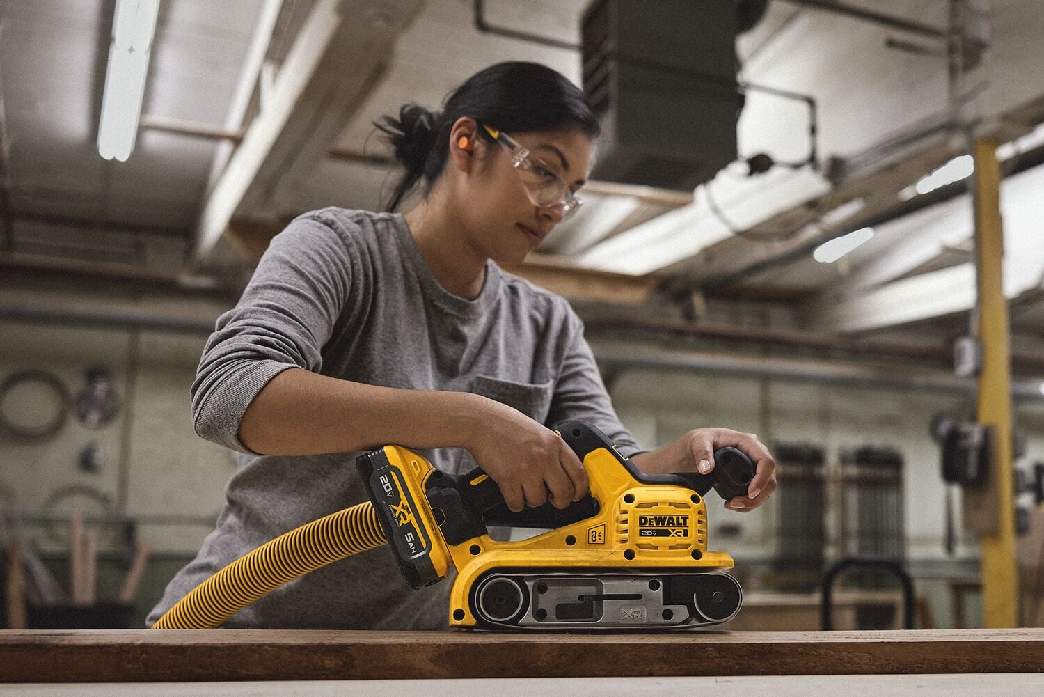 A person using a yellow DEWALT cordless belt sander with a dust extraction hose attached, working on a wooden surface in an indoor workshop.
