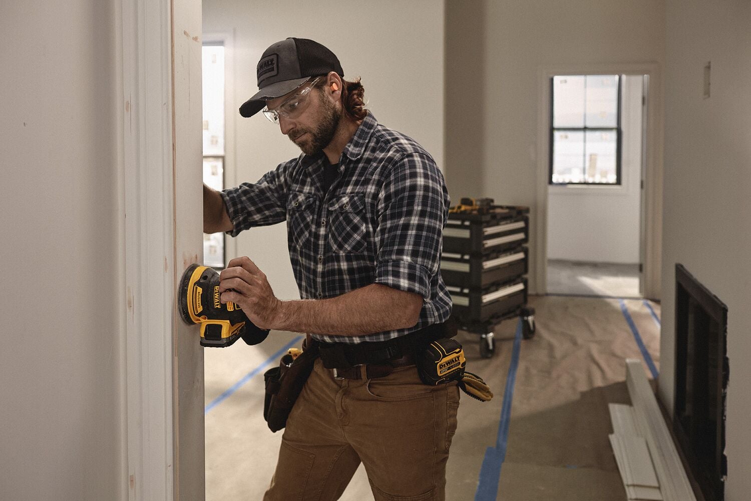 A person uses a DEWALT cordless sander to smooth a wooden doorframe in a construction or renovation setting. The person is wearing work clothes, a tool belt with DEWALT tools, and a cap. Construction materials and equipment are visible in the background.