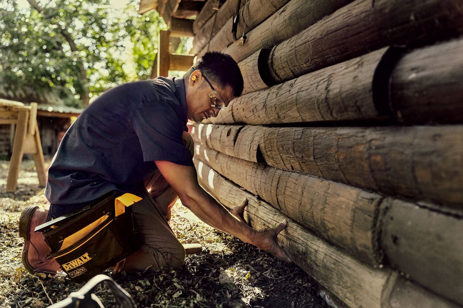 A person wearing a dark shirt and work pants is kneeling on the ground next to a wooden log wall, using their hands to inspect or work on the logs. The person has a DEWALT tool pouch attached to their waist, and natural sunlight is visible in the background.