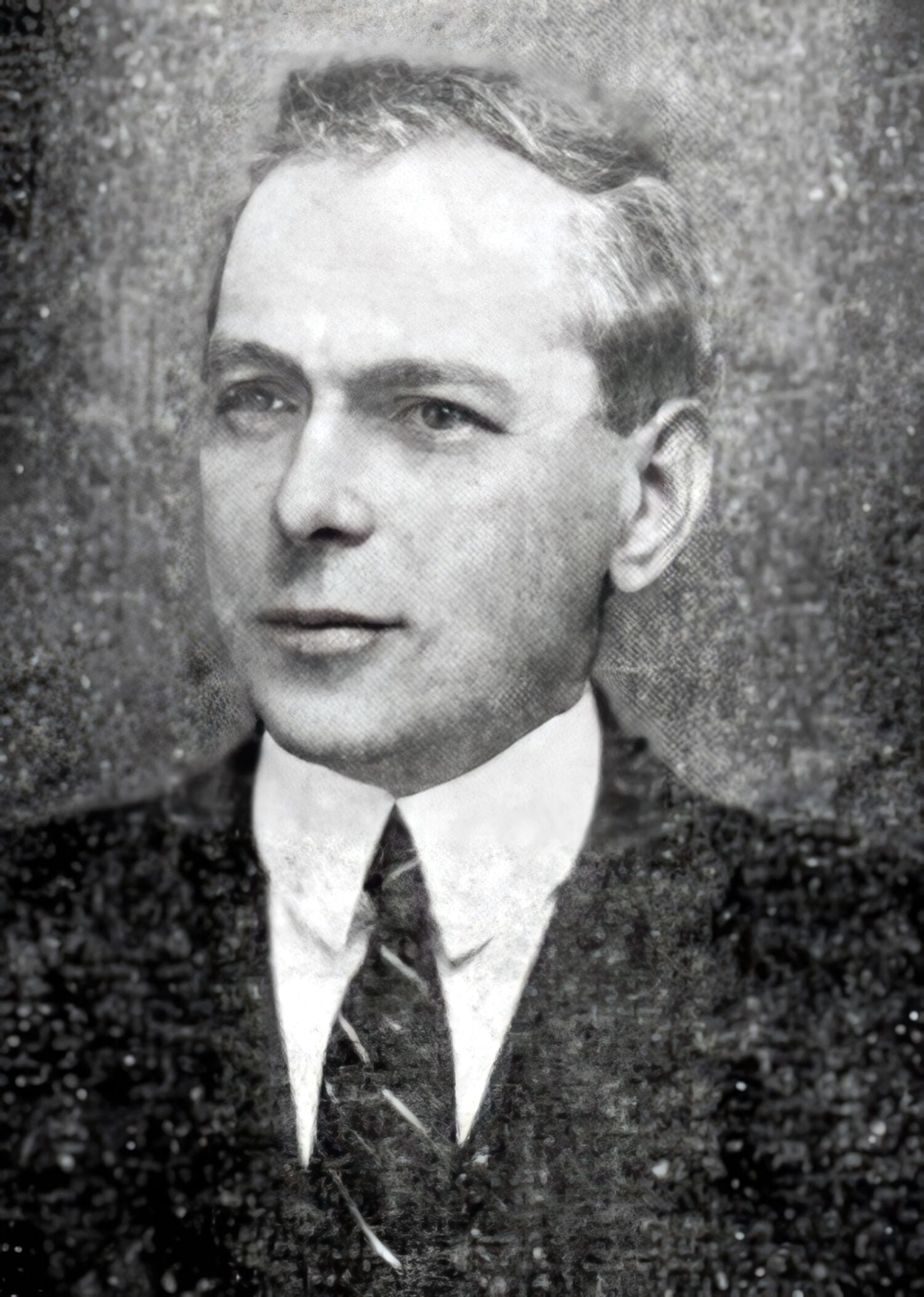 A black and white portrait photograph of a person wearing a suit and tie. The face is obscured by a grey rectangle, making the person unidentifiable.