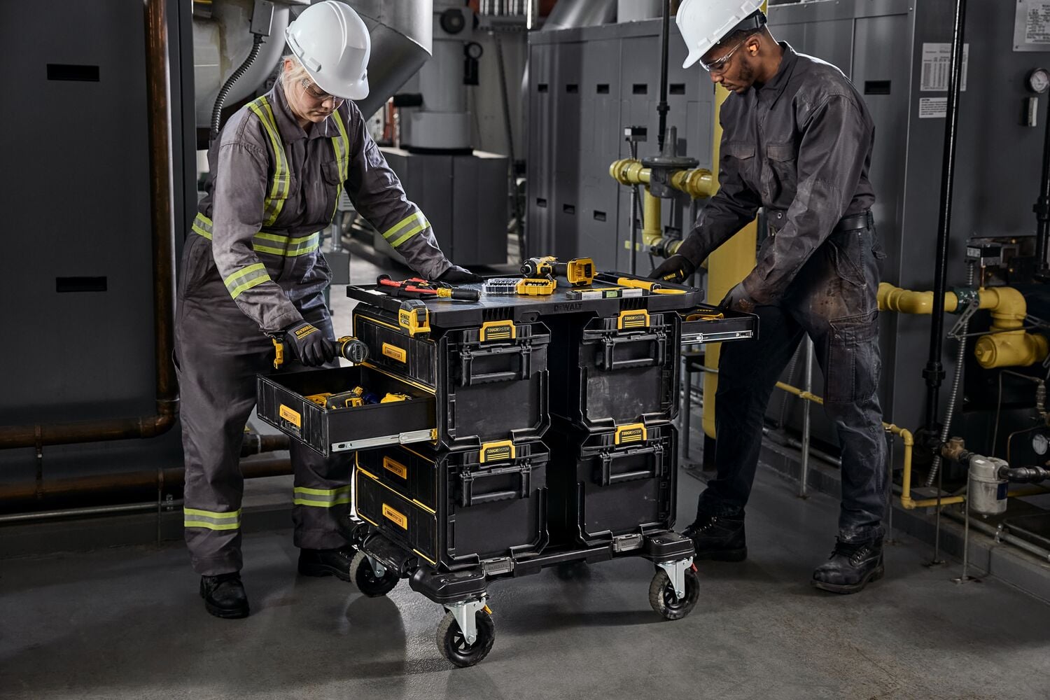 Two workers wearing safety helmets and reflective uniforms are handling a DEWALT rolling tool storage cart with multiple drawers in an industrial setting. Some tools are placed on top of the cart.
