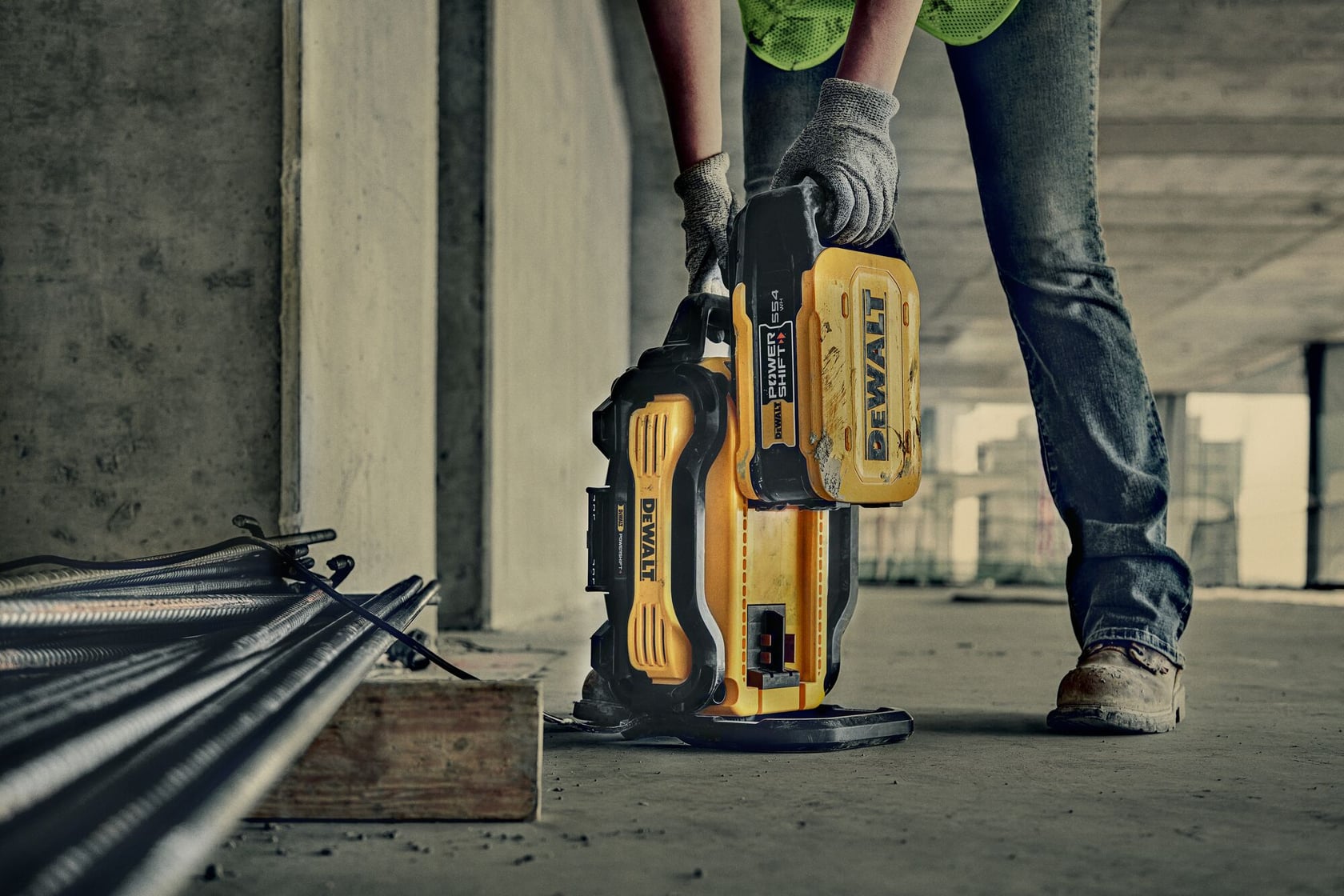 A person wearing work gloves and boots is using a yellow DEWALT portable power supply at a construction site. The device is placed on the ground near metal rods and concrete structures.