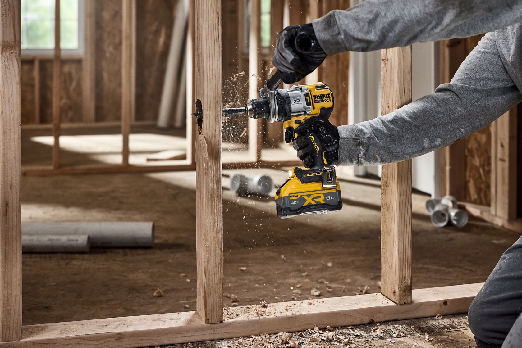 A person using a DEWALT cordless power drill to bore a hole into a wooden frame at a construction site. Wood shavings are visible as the drill operates.