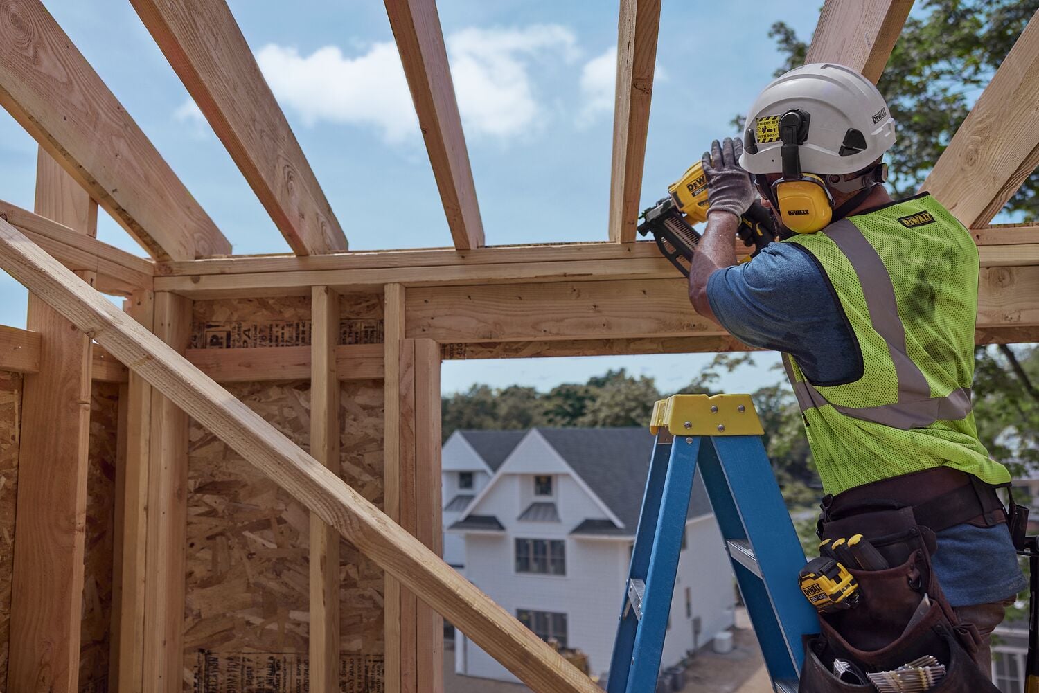 A construction worker wearing a safety helmet and vest is using a DEWALT cordless nail gun to fasten wooden beams while standing on a blue ladder inside a partially built house.