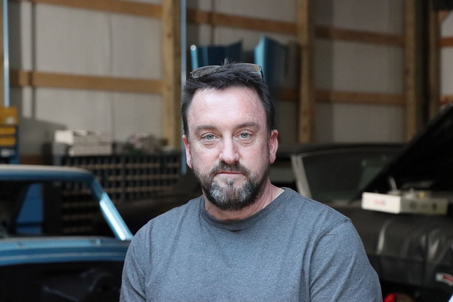A person wearing a gray shirt is standing inside a garage or workshop. The face is blurred for privacy. There are cars and shelving visible in the background.