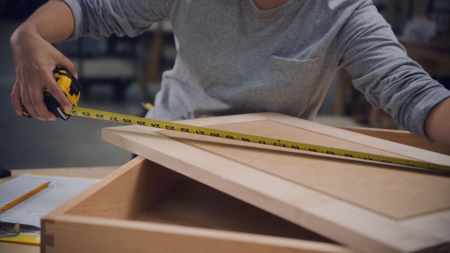 A person using a DEWALT yellow tape measure to measure a wooden cabinet door in a workshop.