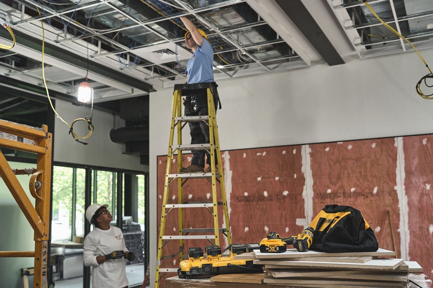 A construction scene showing a worker on a yellow ladder installing cables in a ceiling. In the foreground, DEWALT power tools and a black and yellow tool bag are placed on a stack of wooden boards. Another person stands on the ground below, looking up. The environment is indoors with exposed ceiling infrastructure and unfinished walls.