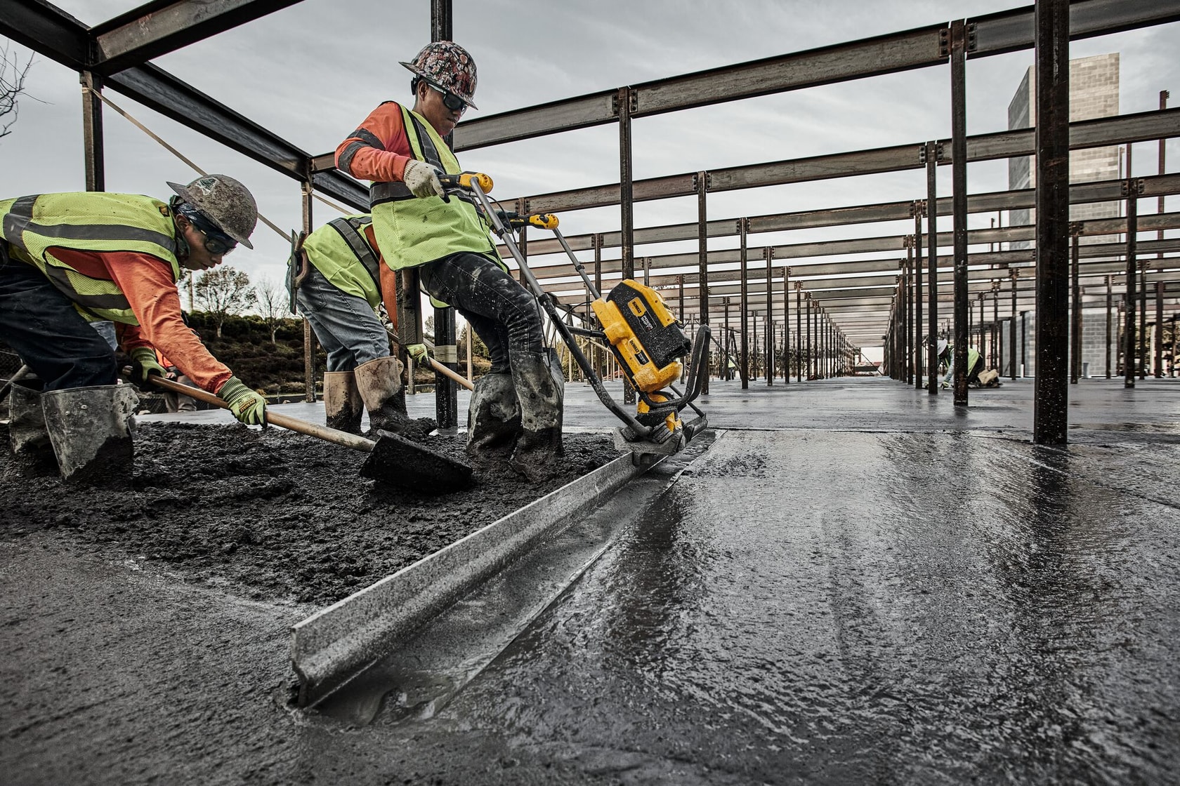 Workers wearing safety vests and helmets are leveling concrete at a construction site using a DEWALT power tool. The area is structured with steel beams, and the ground appears freshly poured and smoothed.