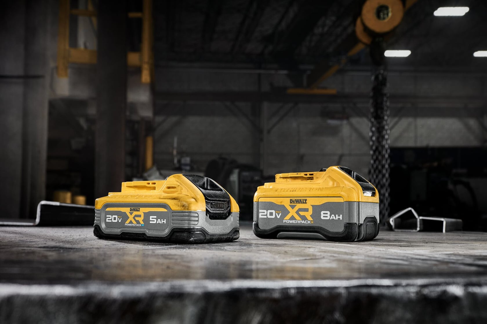 Two DEWALT 20V XR lithium-ion batteries are placed side by side on an industrial workbench. One is labeled 5Ah and the other is labeled 8Ah, both featuring yellow and black casing. The workshop setting is visible in the background.