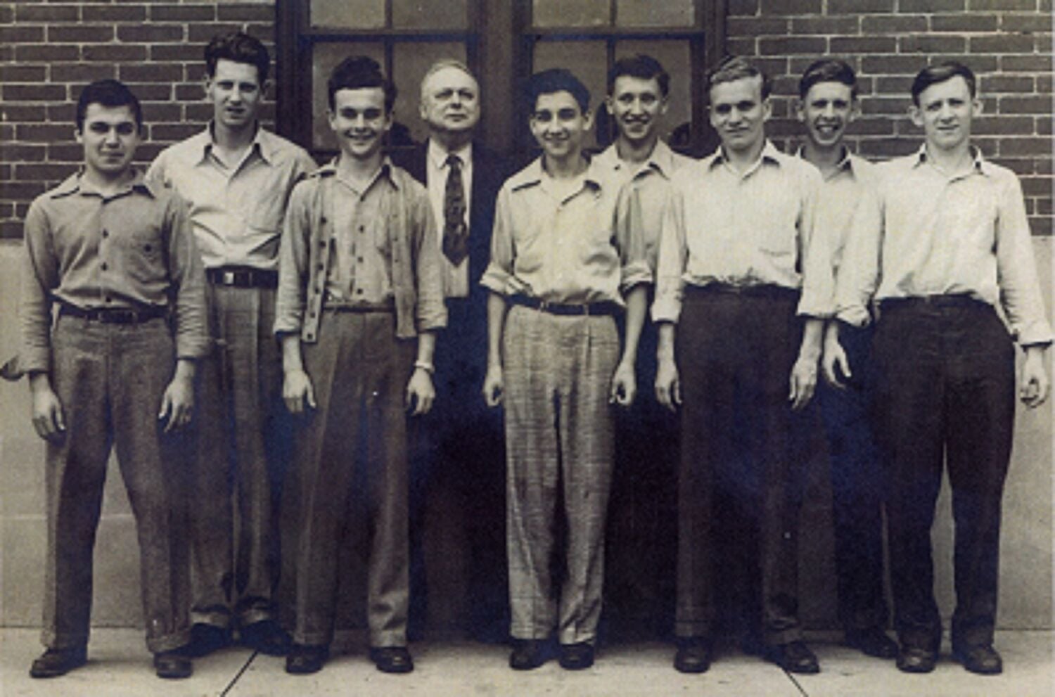 A vintage black and white photo showing a group of eight men standing in front of a brick building with their faces intentionally blurred.
