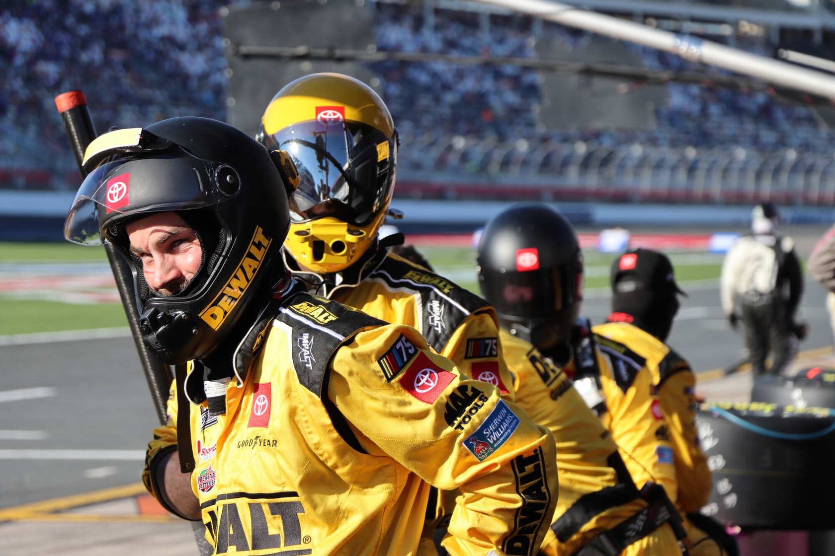 Several pit crew members wearing yellow DEWALT-branded uniforms and helmets are positioned by a race car at a motorsport event. The crew appears ready for action, with blurred faces for privacy. The background shows a racetrack and spectators.