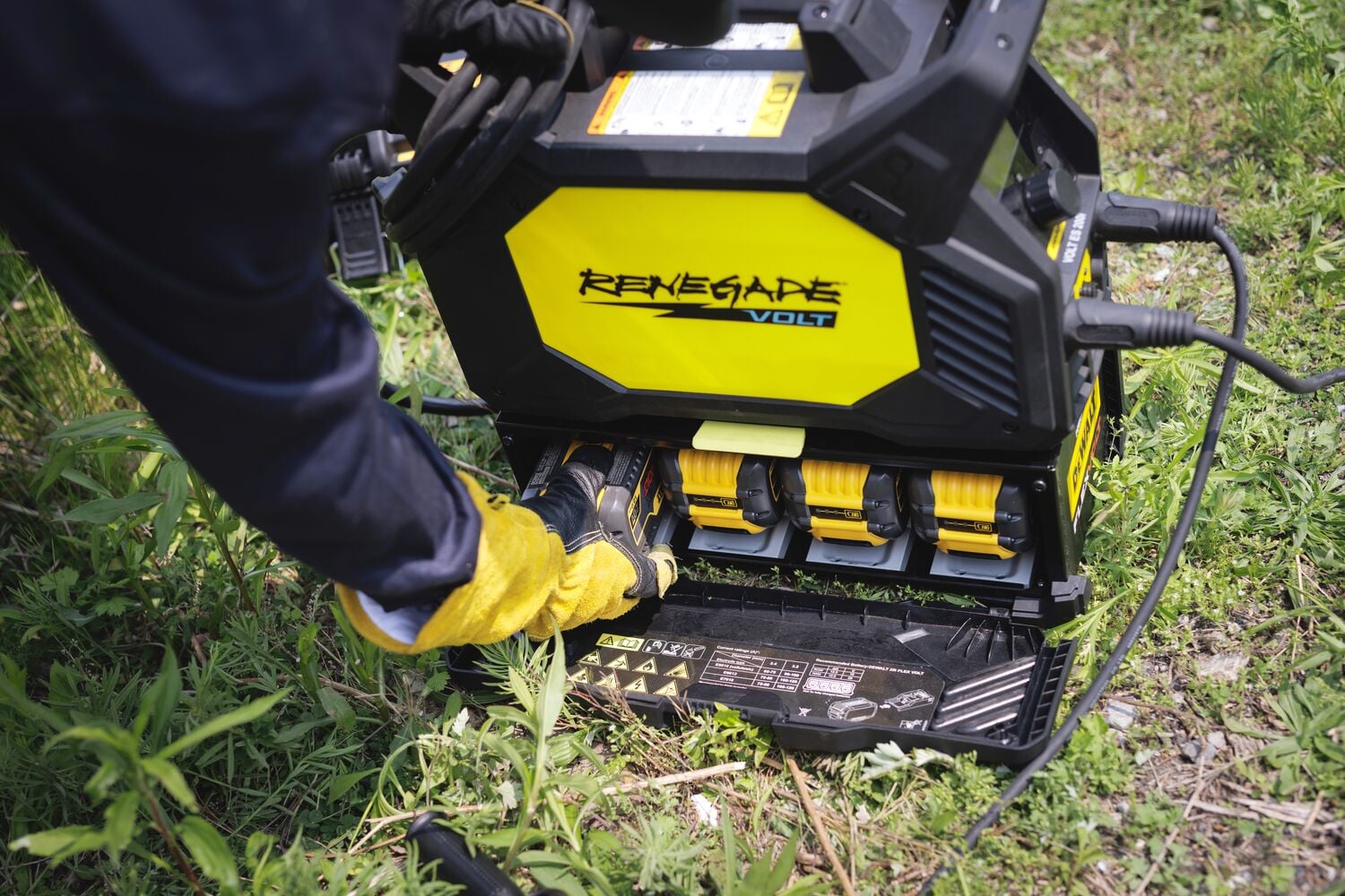 A person wearing gloves is inserting a DEWALT battery into a yellow and black Renegade Volt device outdoors on grassy ground.