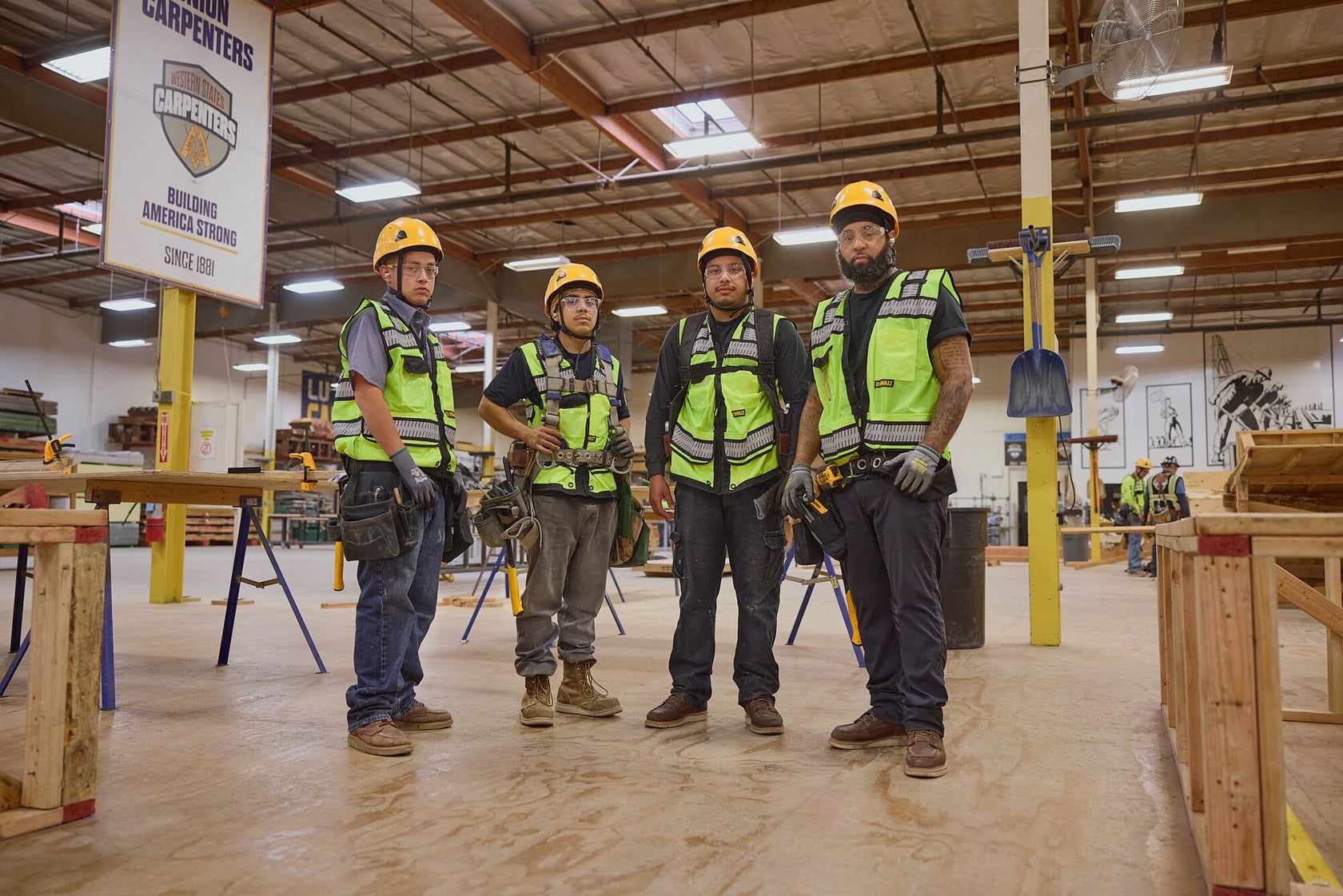 Four construction workers wearing safety helmets and green reflective vests stand together inside a spacious workshop. The workshop features woodworking tables, tools, and equipment. The workers' faces are blurred for privacy. A sign in the background mentions carpenters and building America strong.