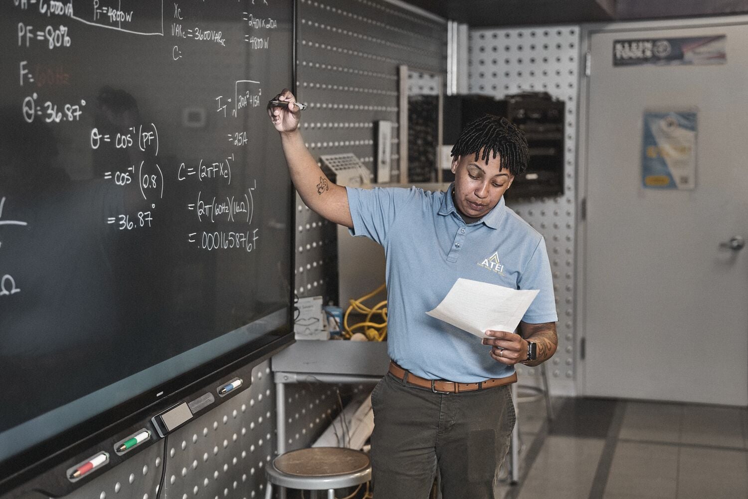 A person with a blurred face wearing a light blue polo shirt stands in a classroom and writes electrical equations on a blackboard, holding a piece of paper in the other hand.