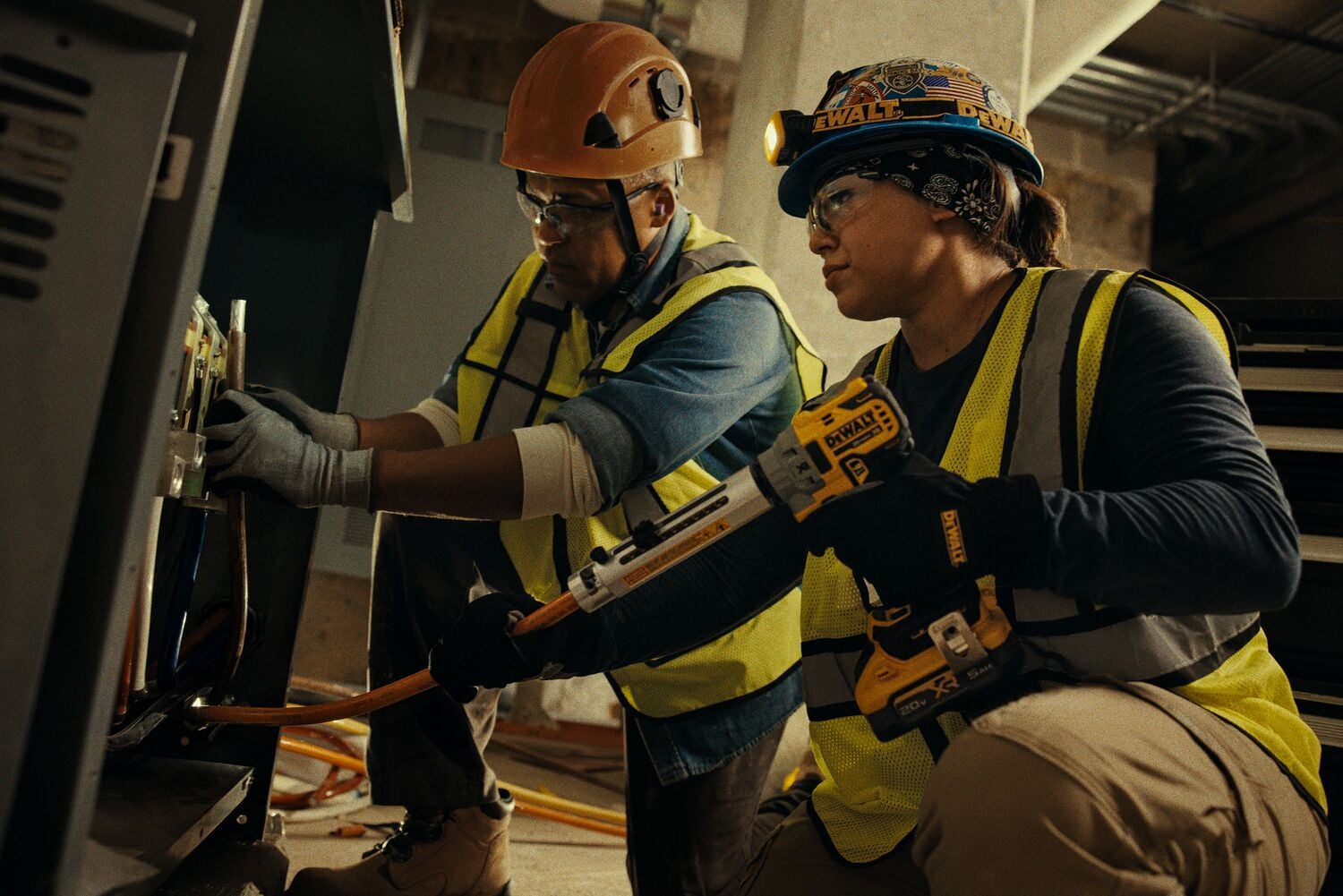 Two construction workers wearing safety vests and helmets are using a DEWALT caulking gun tool. They are working near an industrial electrical panel in an indoor environment.
